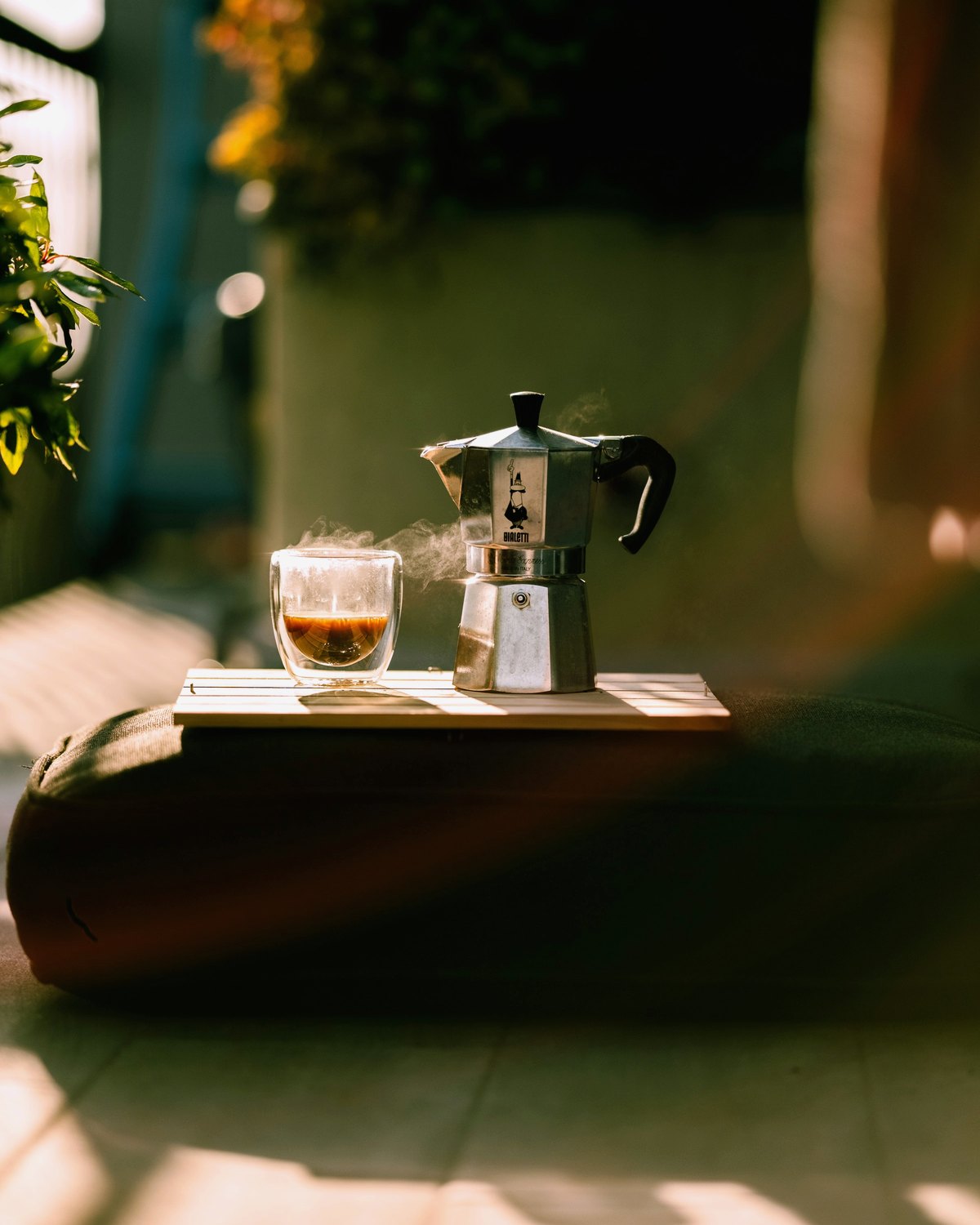 A glass of freshly brewed coffee steaming beside a moka pot in the morning sun